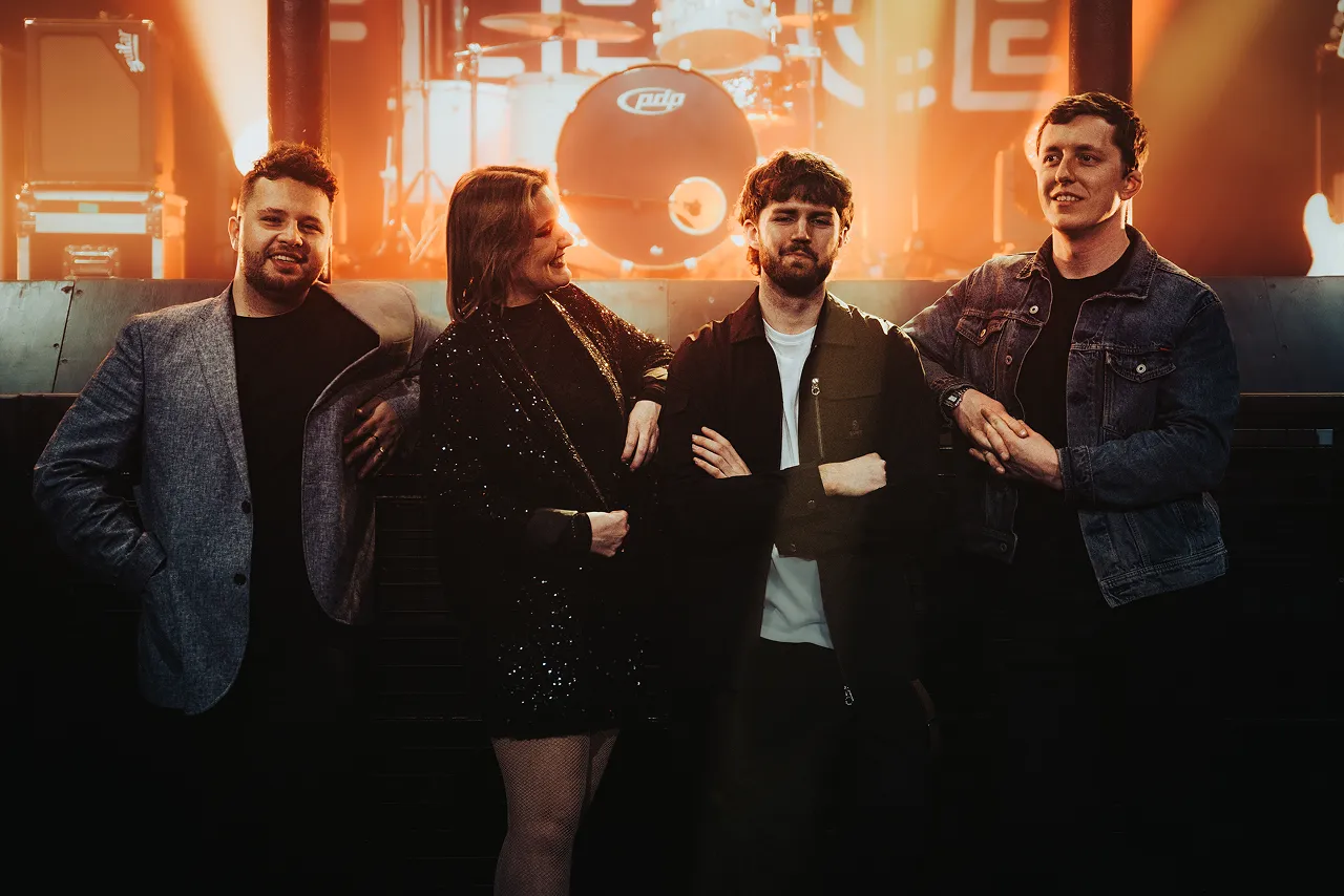 Four young adults posing on a stage with drum set in background under warm lighting.